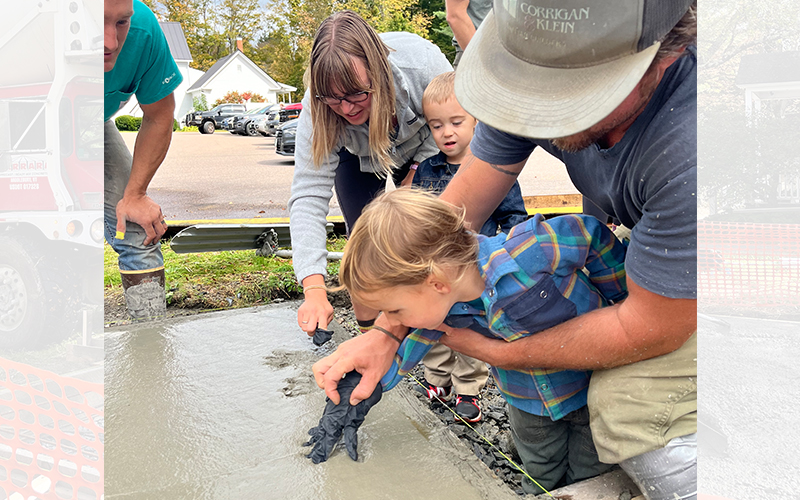 E. Adam Wood (Richmond) Jeremiah Kemp (Warren) observe as Caitlin Ennis and son Duncan (Fayston) and Rhys Corrigan and father Tayloir Ciorrigan (Warren) place handprints in the fresh concrete. Photo: Devin Klein Corrigan 